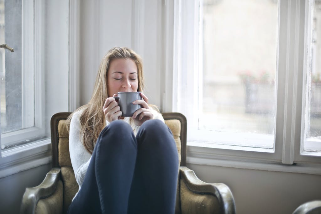 Woman enjoying a warm drink in a cozy armchair by a window, embracing relaxation.